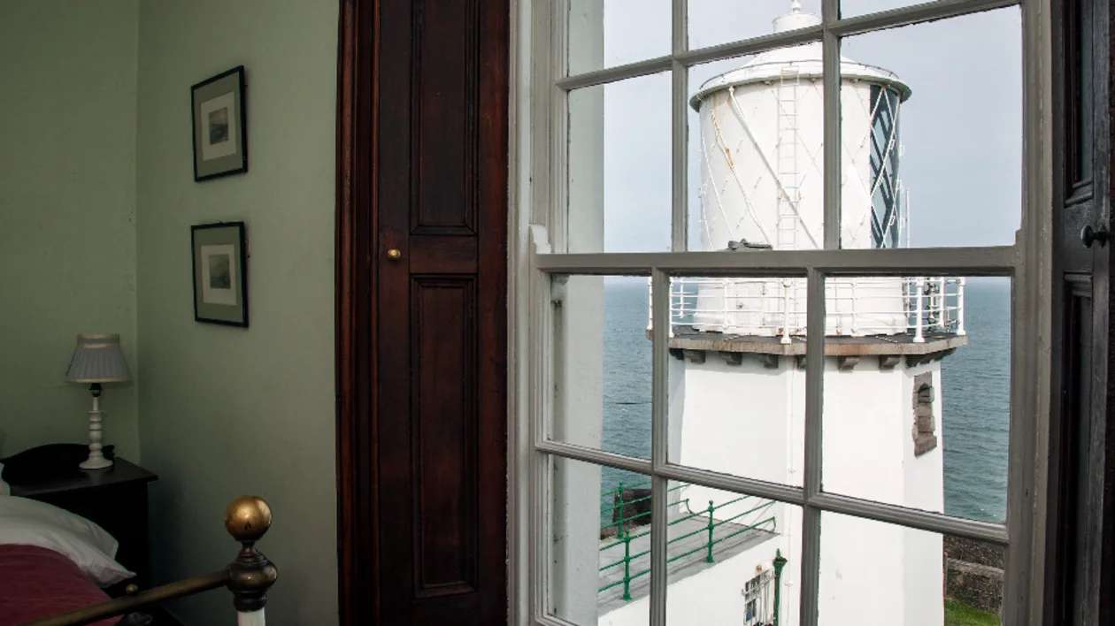 A view of a white lighthouse seen through a window from a cosy room.