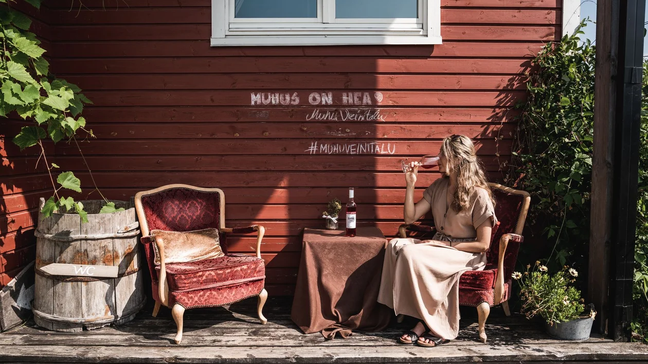 Woman enjoying a glass of rosé at Muhu Winehouse in Estonia, sitting by a rustic red wooden house.