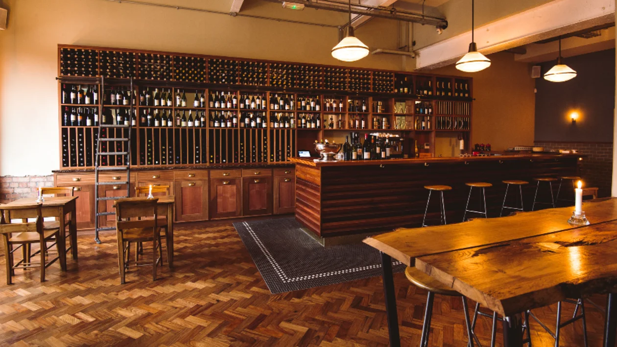 A cozy wine bar interior featuring a wooden counter, shelves filled with bottles, and rustic tables under warm lighting.