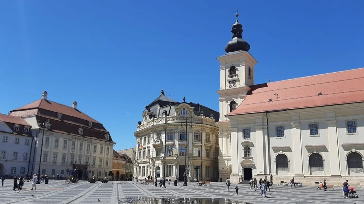 A sunlit square featuring historic buildings with varying architectural styles, bustling with people under a clear blue sky.