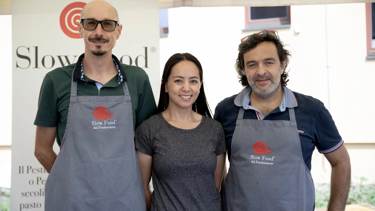 Three people smiling at a Slow Food del Pordenonese event, wearing aprons and standing in front of the organisation’s banner.