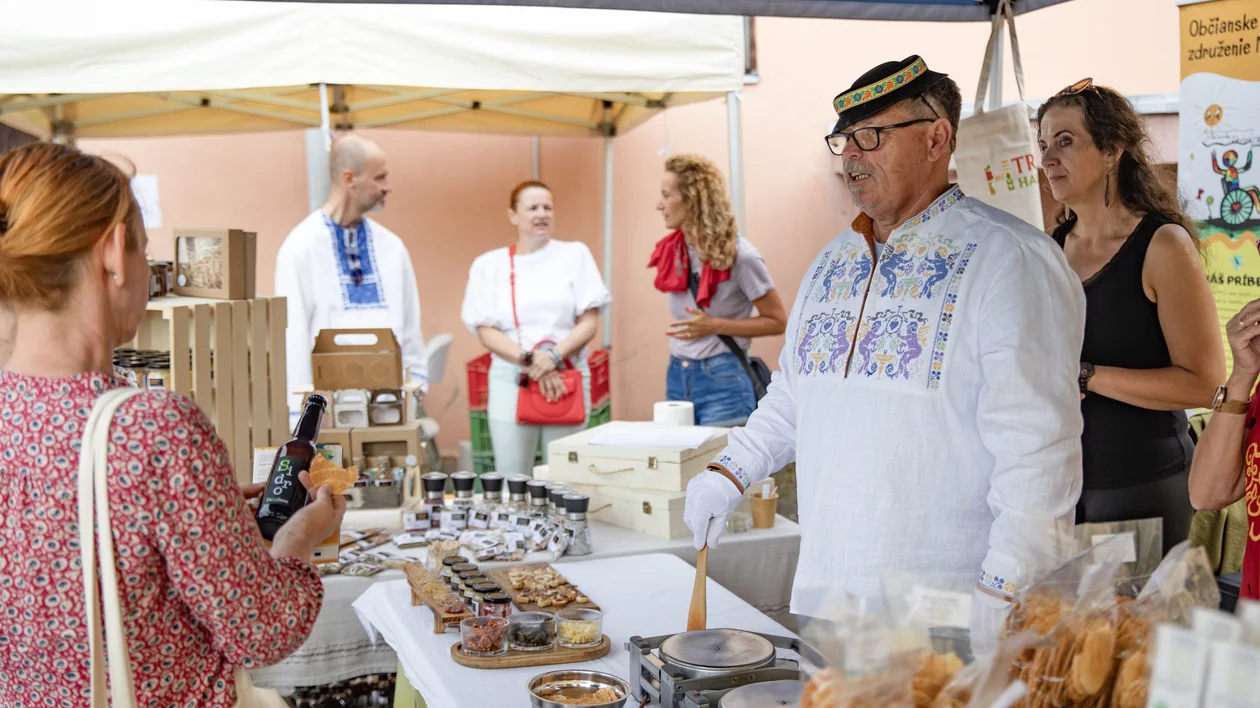 Man in traditional embroidered shirt serving customers at a farmers’ market stall with local products in Bátovce, Slovakia."