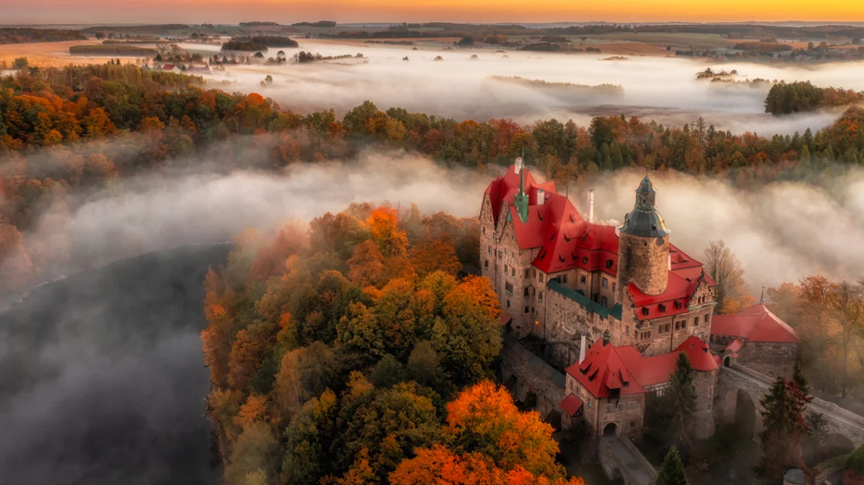 Elevated view of Czocha Castle surrounded by fall foliage in orange and green tones beneath a partly cloudy sky.