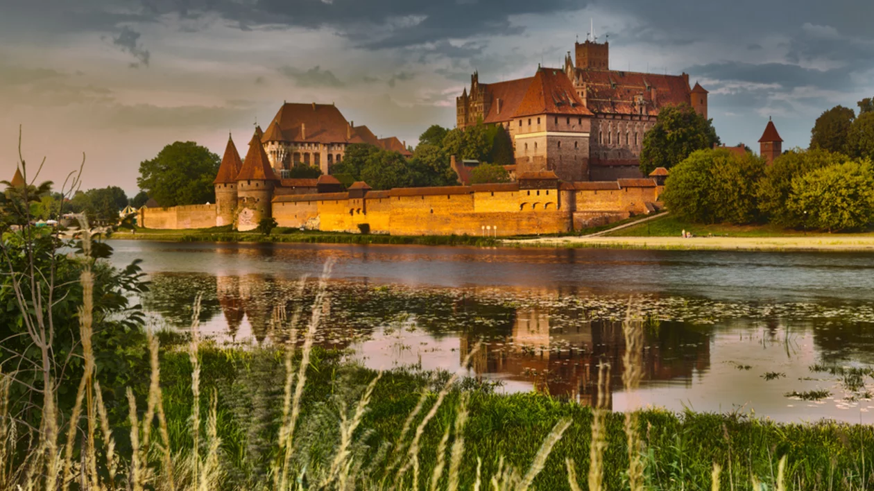 Marlborough Castle seen across a river, surrounded by lush green landscape and reflected in the water beneath clouds.