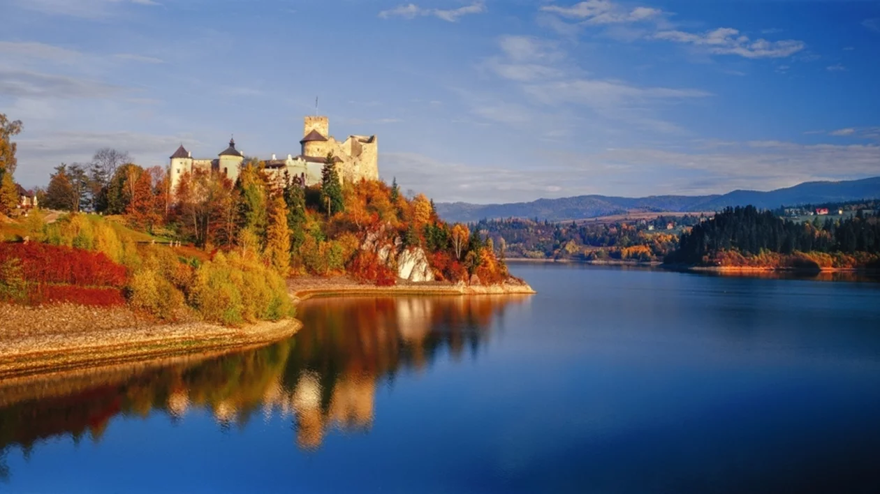 Autumn landscape at Niedzica Castle with colorful trees in green, orange, and red, mountainous backdrop, and a river winding below the hill beneath a clear sky.