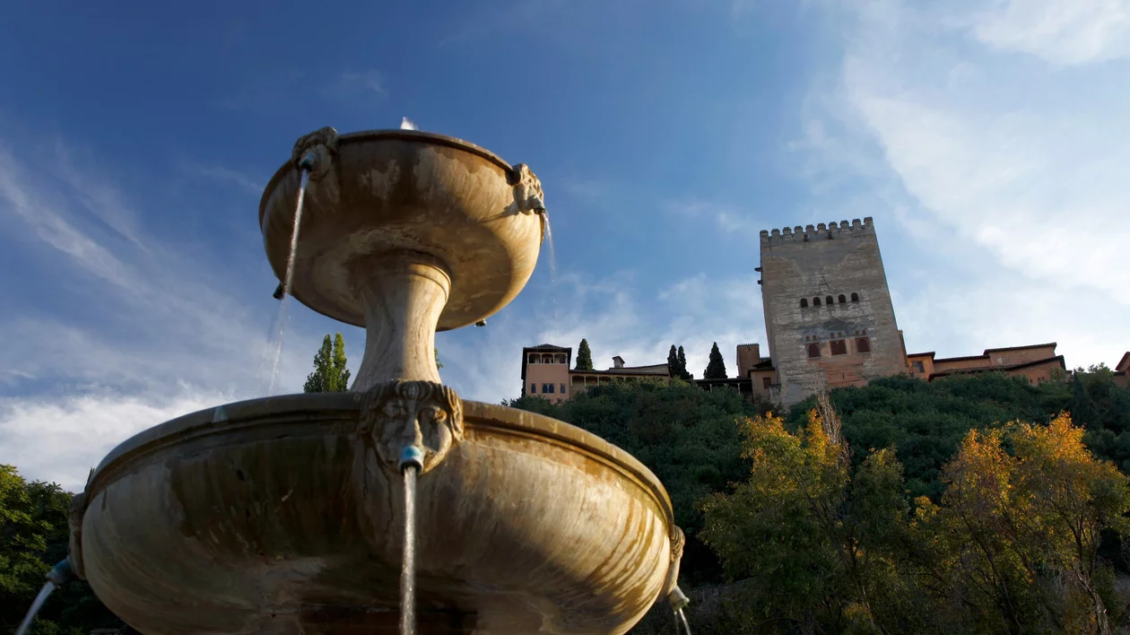 Photo taken from below featuring a fountain in the foreground with water flowing from its sculptures, the Paseo de los Tristes behind it, the castle in the background, green trees, and a blue sky above, illuminated by sunlight.