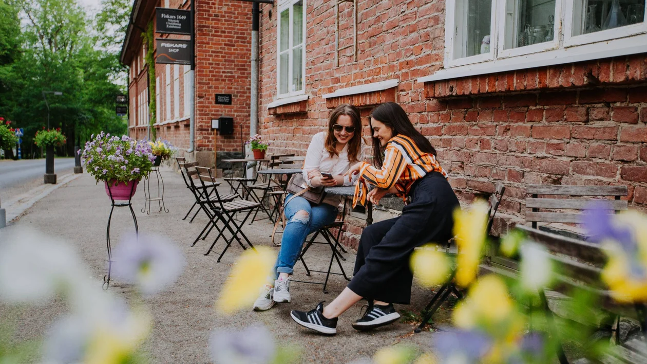 Friends sitting in a café in Southern Finland, smiling and looking at a phone, surrounded by flowers with green trees in the background.