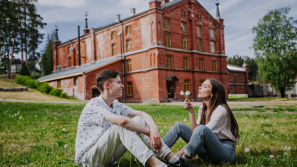 Couple sitting in front of the Kouvola board mill, a UNESCO World Heritage site, blowing a dandelion on a summer day, surrounded by green scenery.
