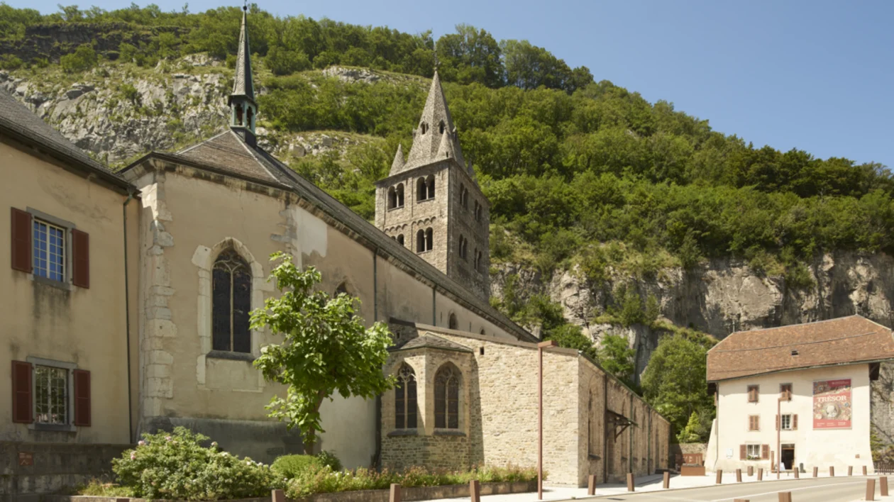 Historic stone church with a bell tower surrounded by green hills on a sunny day in Saint-Ursanne, Switzerland.