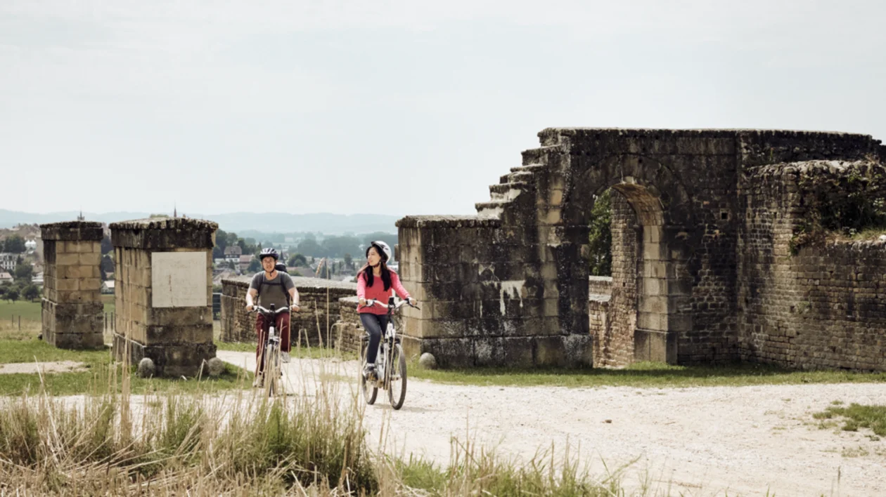 Cyclists riding along the ancient Via Romana surrounded by historic ruins and green landscape on a sunny day.