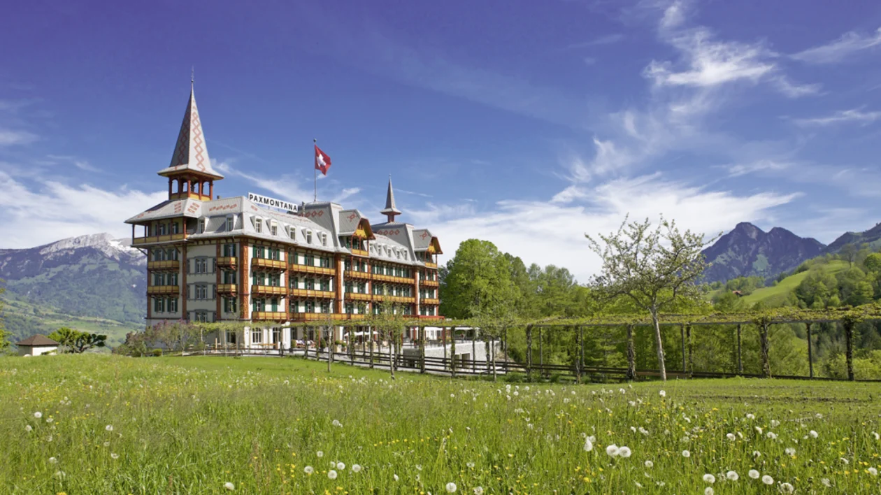 View of a hotel in the green landscape of Kerns village, with a Swiss flag and dandelions under a sunny sky with light clouds.