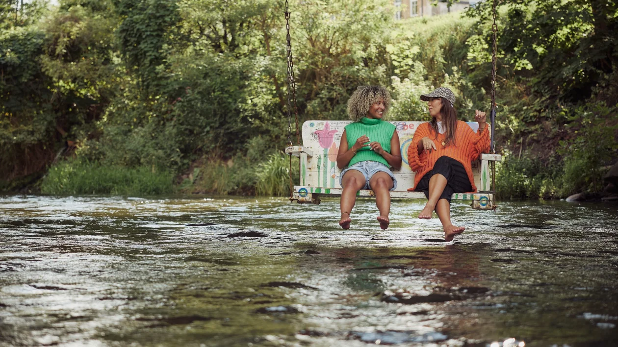 Two friends sitting barefoot on a swing over the Vilnia River in Vilnius, laughing and enjoying a summer day surrounded by greenery.