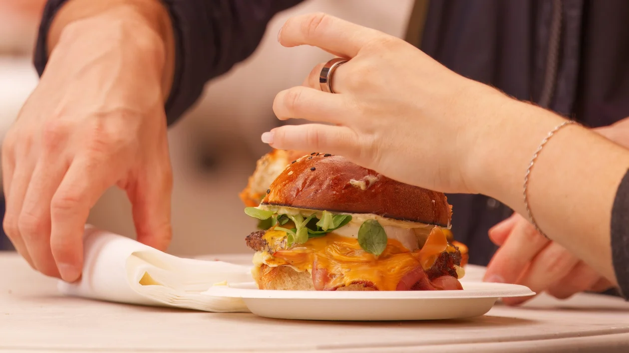 Close-up of two people sharing a gourmet burger with melted cheese, fresh greens, and a brioche bun at an outdoor food market.