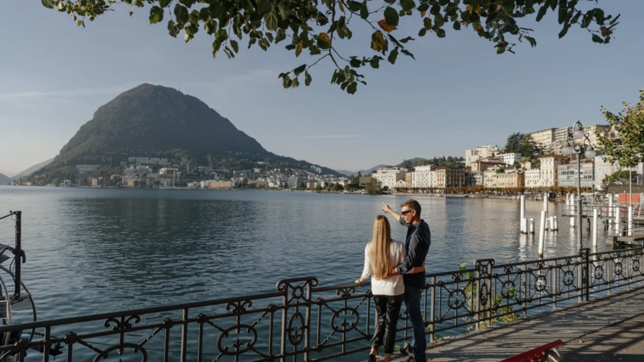 Panoramic view of Lugano, Switzerland — the lakeside city with terracotta rooftops, Lake Lugano, and surrounding green hills.