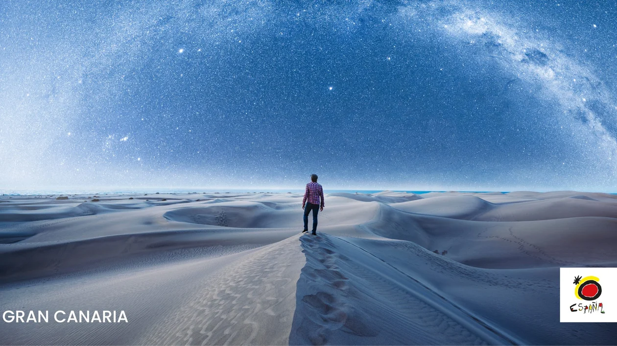 Person standing on a sand dune in Gran Canaria under a clear, star-filled night sky stretching across the desert landscape.