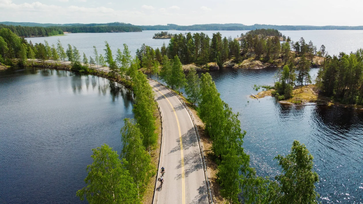 Cyclist riding a bicycle on a forested trail along the shores of Lake Saimaa in Finland, with calm water and lush green trees in the background.