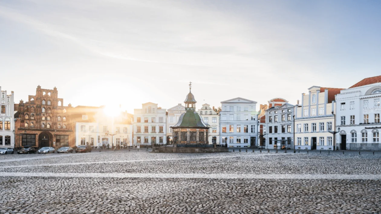 Historic European town square paved with cobblestones, featuring a central fountain and tall, gabled buildings under a bright sun.