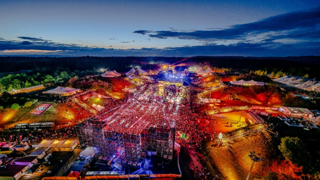 Aerial night view of a massive, crowded outdoor music festival with bright stage lighting and multiple large tent structures.