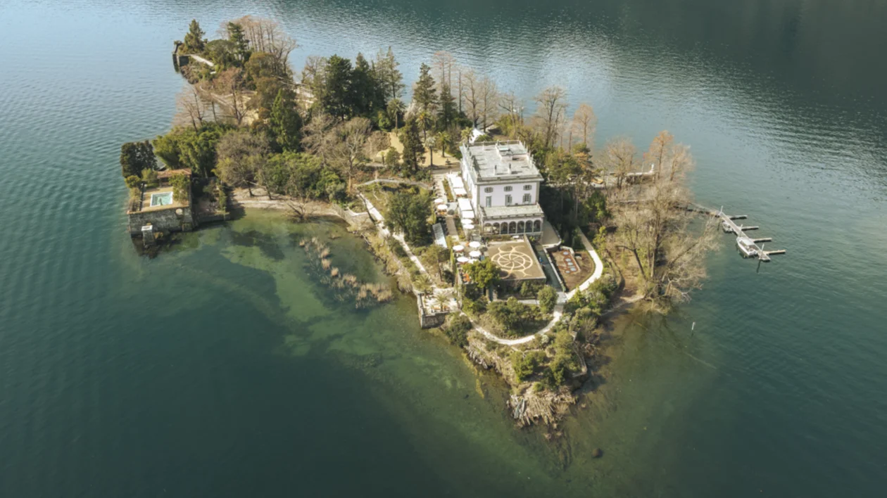 Small island with a large house surrounded by clear blue water and green trees.