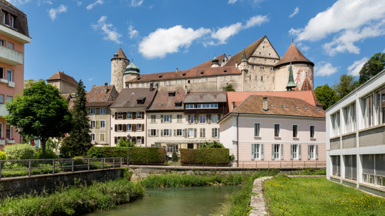 Large historic castle on a hill, surrounded by houses and a stream under a blue sky.