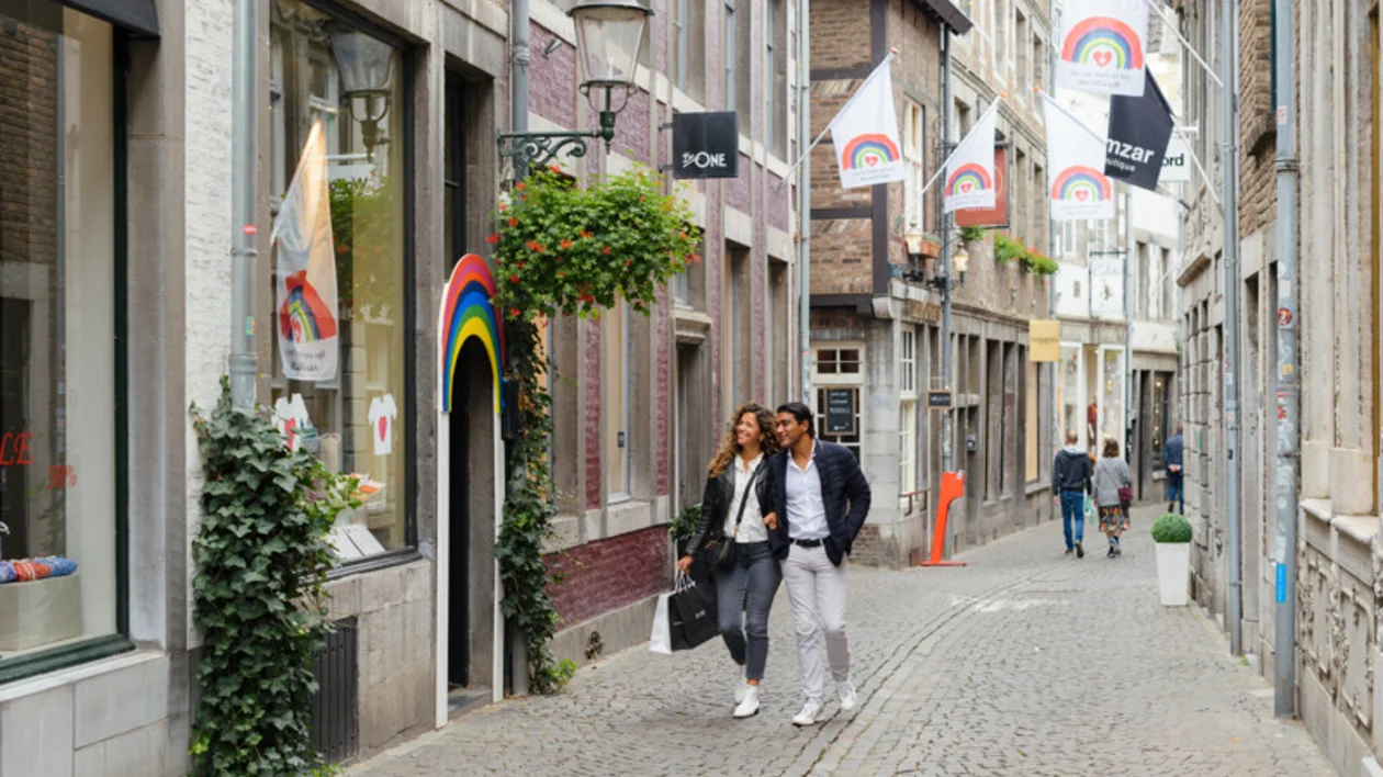Couple walking down a cobblestone street lined with shops and rainbow flags.