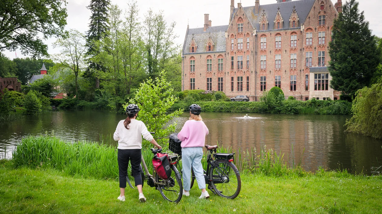 Couple with bicycles by a lake facing a historic castle in Belgium.
