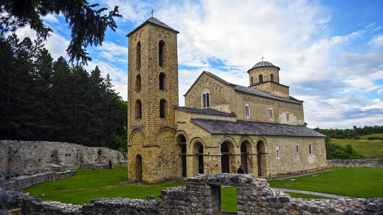 Stone church with arched windows and tower, surrounded by grass and trees under a blue sky.