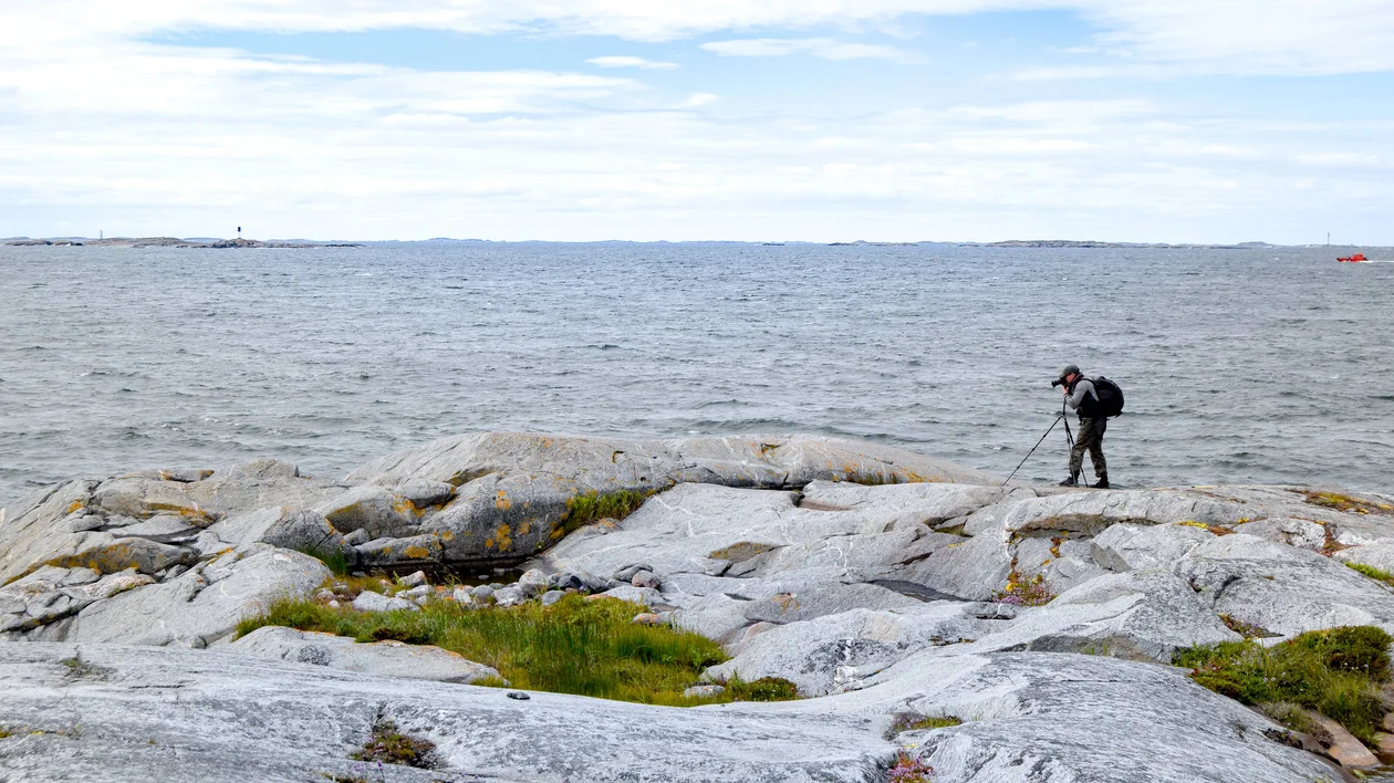 Person taking photos on rocky shore by the sea under a partly cloudy sky.