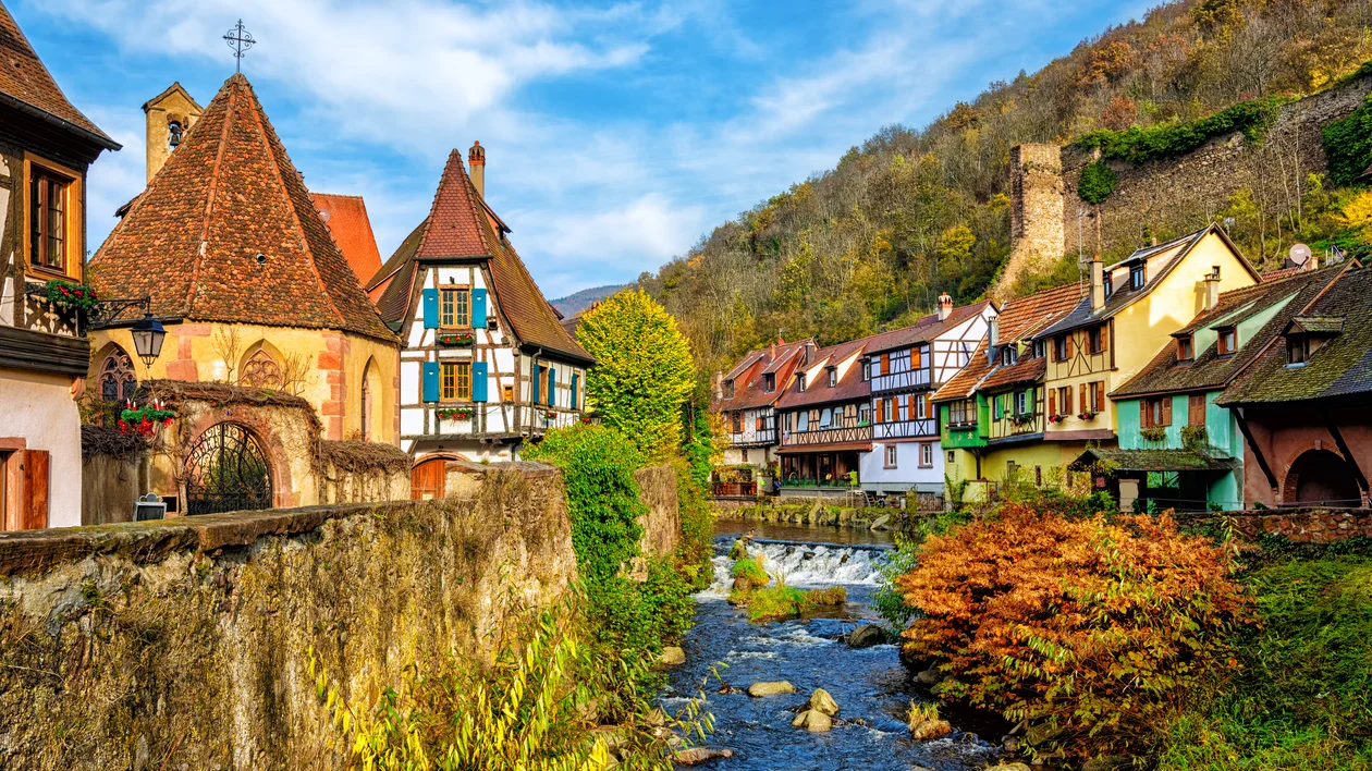 Colorful houses line a river in a quaint village with a hillside and castle ruins in the background.