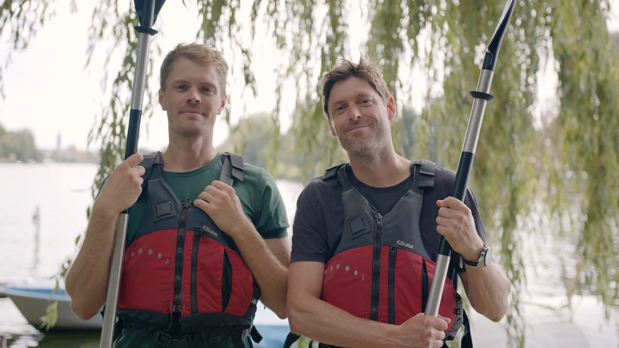 Two men with paddles wearing red life jackets, standing by a lake under willow trees.