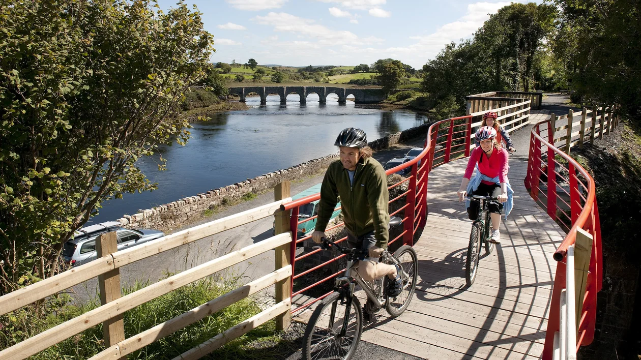 Two people riding bikes on a riverside path with a bridge in the background.