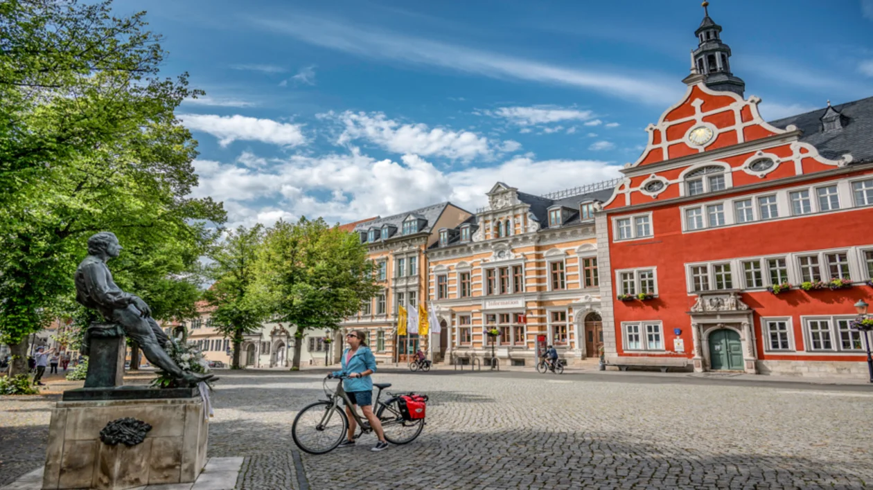 Cyclist passing statue in a cobblestone square with colorful historic buildings and blue sky.
