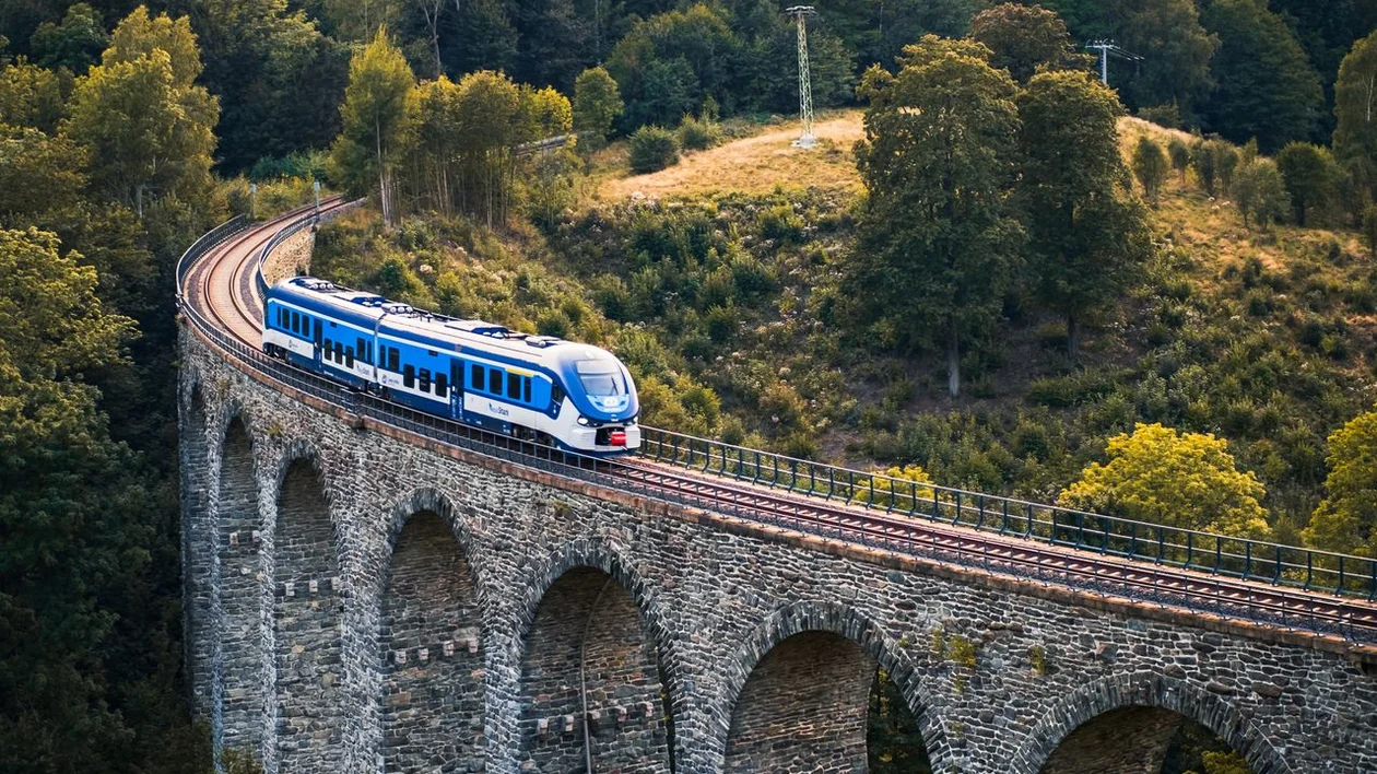 Blue train crossing a stone viaduct bridge in a lush, green hilly landscape.