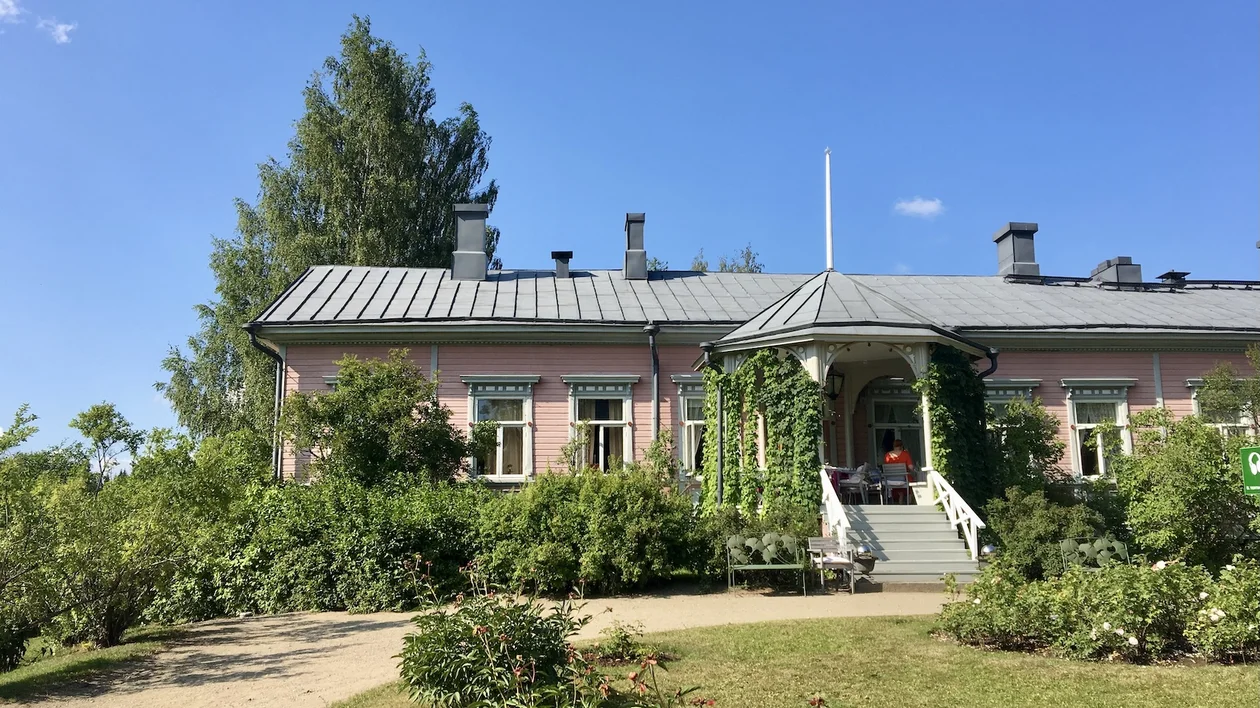 Pink country house with a gray roof, front steps, and lush green garden under blue sky.