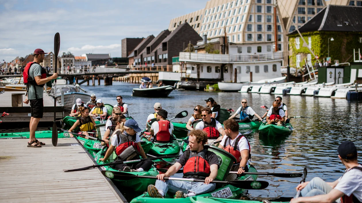 Group of people in kayaks on a canal, listening to an instructor by the dock.