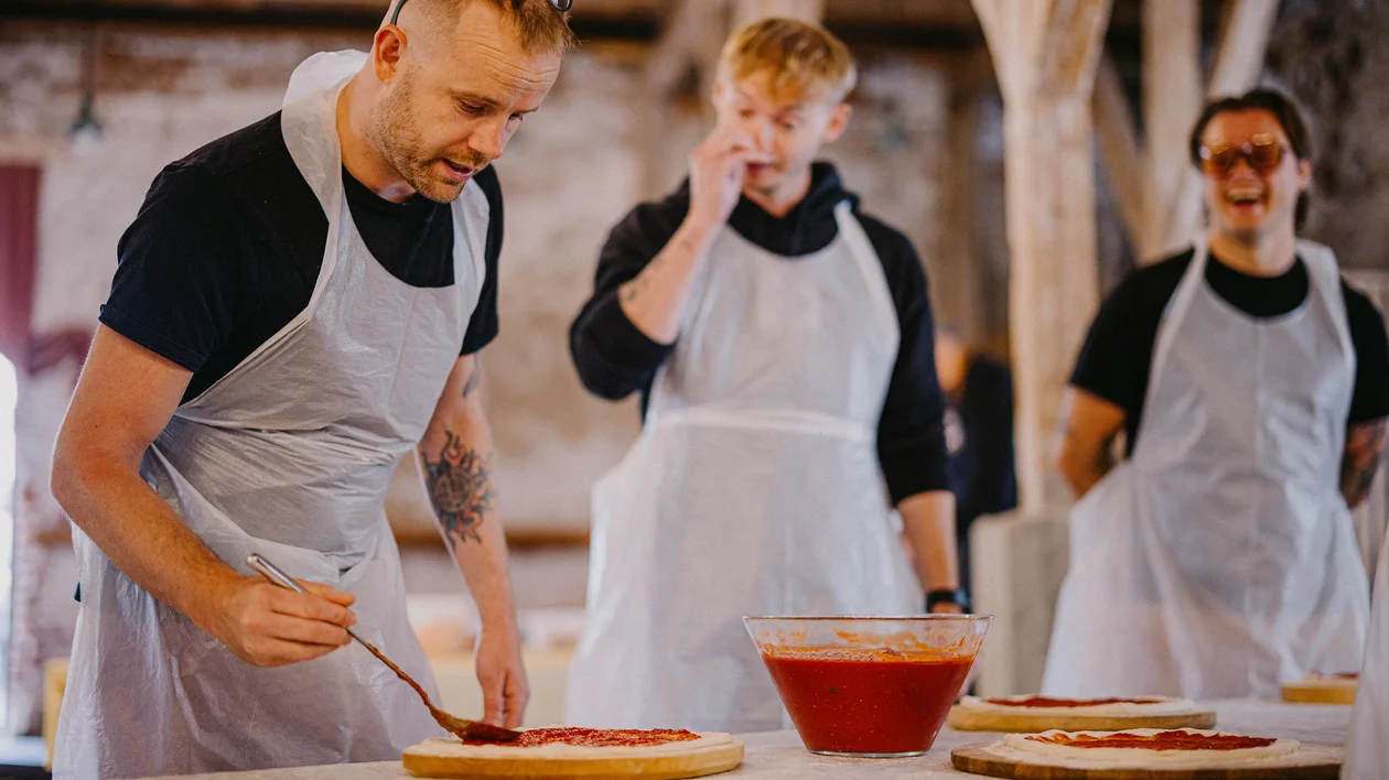 Man spreading tomato sauce on pizza dough, two others smiling in the background.