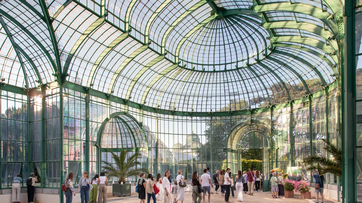 People walking inside a large greenhouse with a glass and metal arched ceiling.