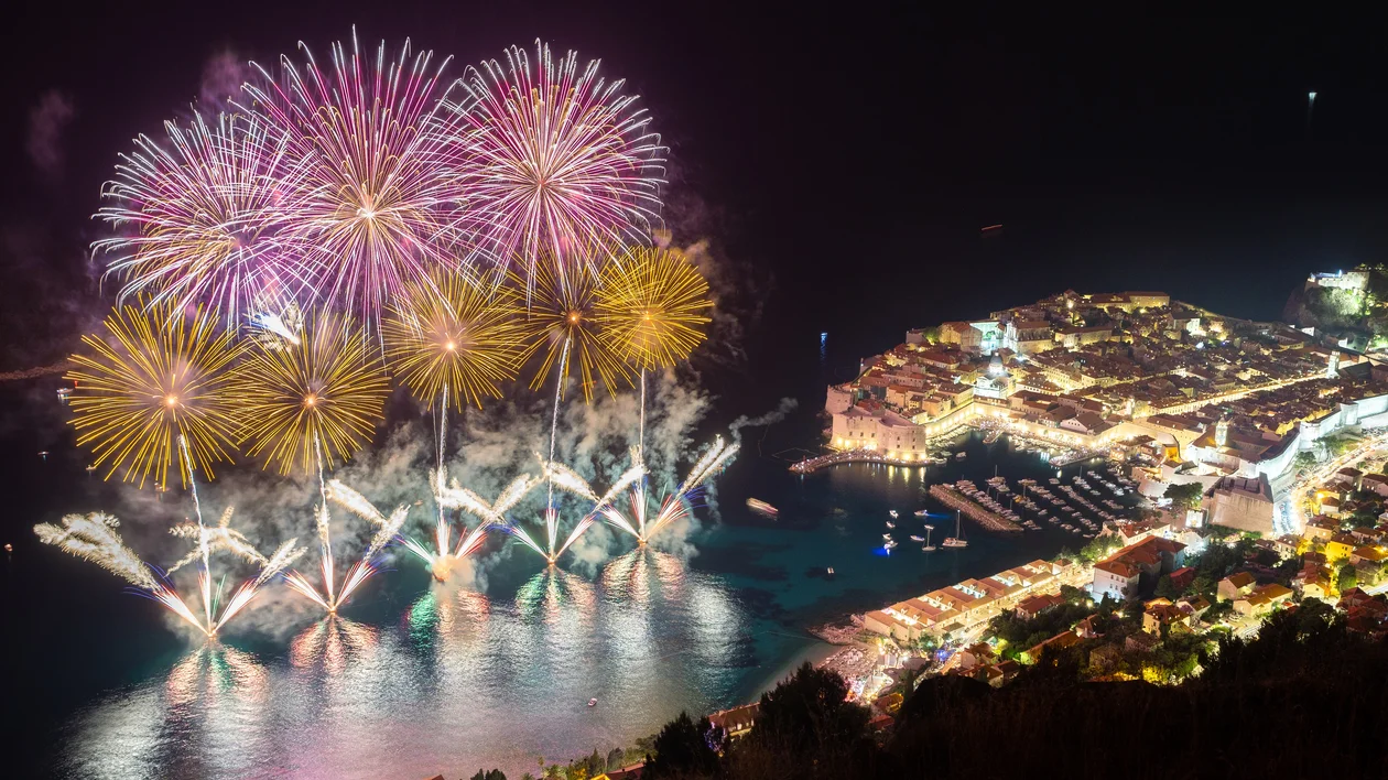 Colorful fireworks erupt over a brightly lit coastal town at night.