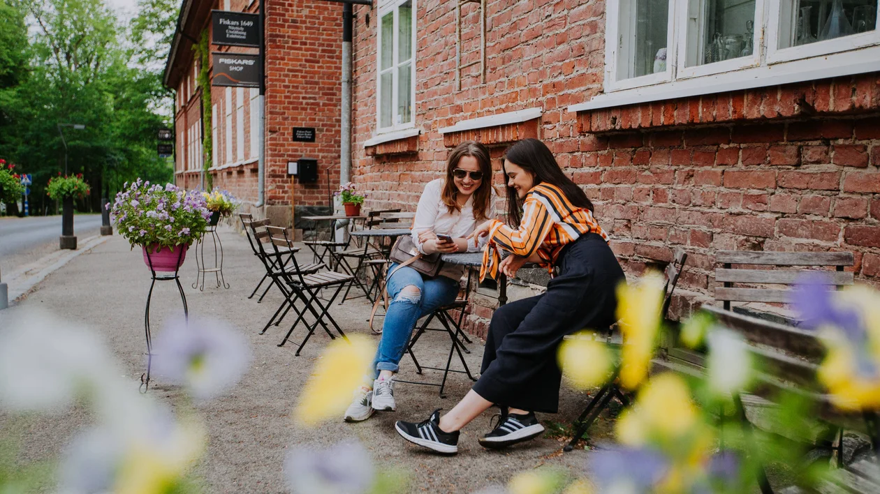 Two women sitting on a bench outside a brick café, smiling at a phone.