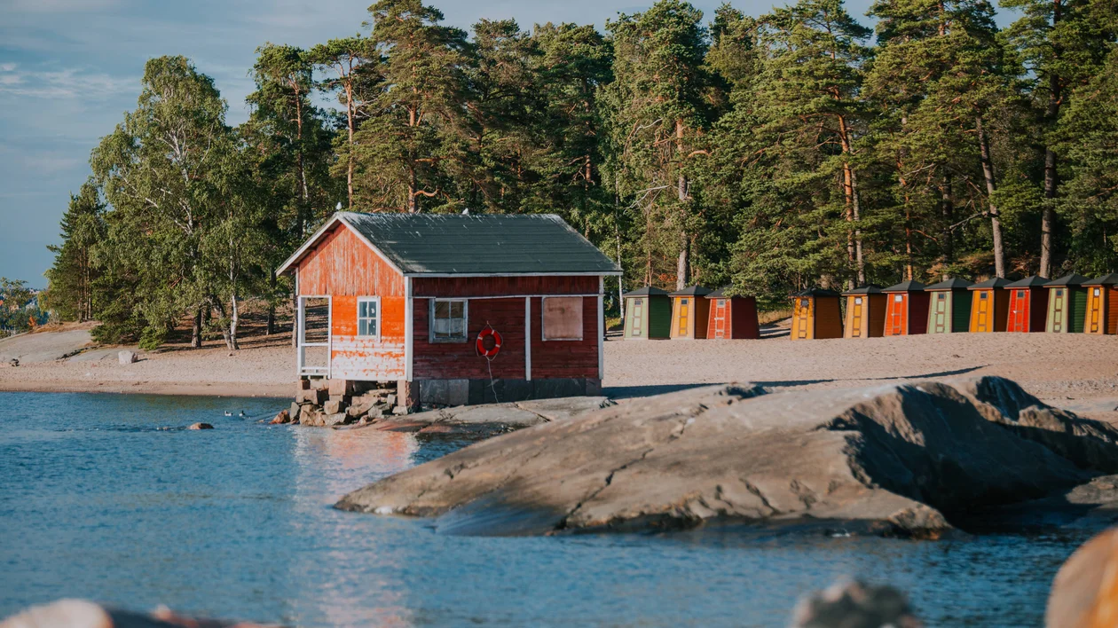 Red cabin by the water, rocky shore, pine trees, and colorful beach huts in the background.