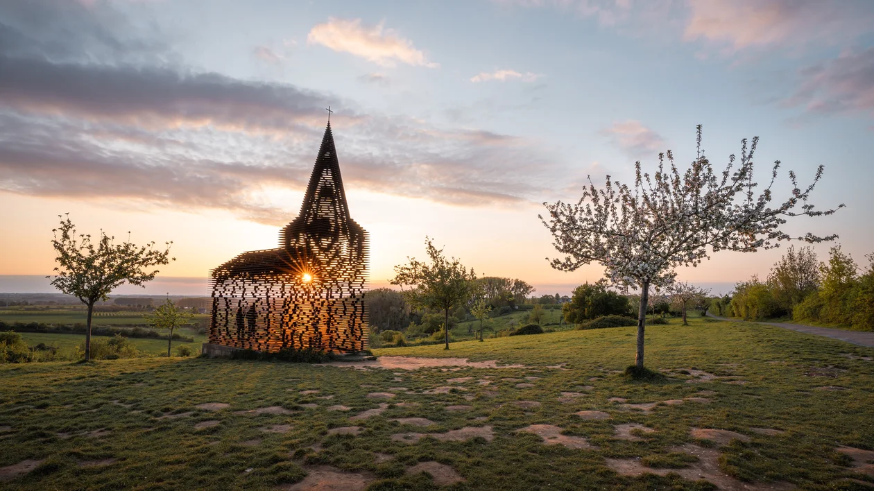 Sunset behind a transparent church sculpture on a grassy hill with scattered trees.