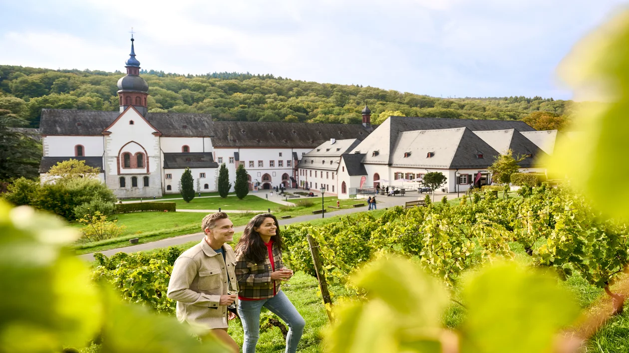 Two people walk through a vineyard near a large white historic building with a church tower.