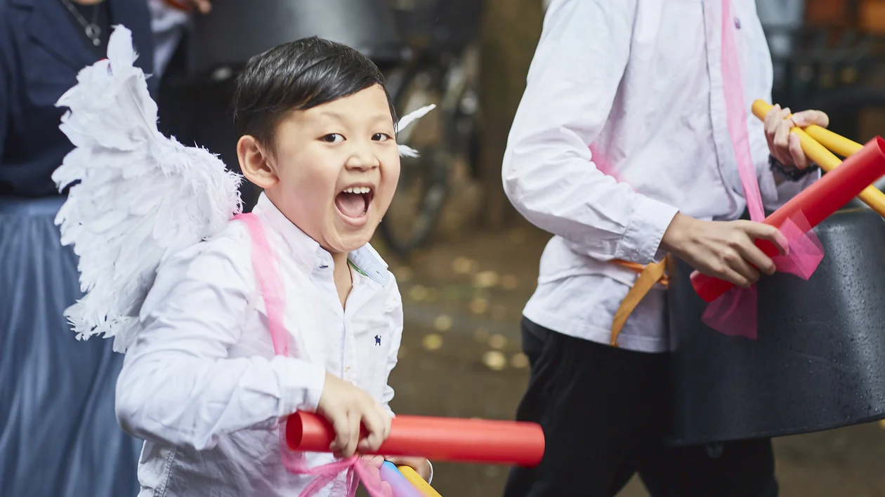 Smiling boy in white shirt and angel wings holding colorful sticks at a parade.