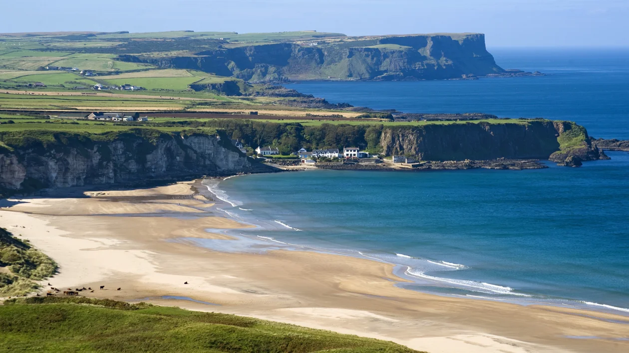 Sandy beach with blue waves, green cliffs, and rolling countryside under a clear sky.