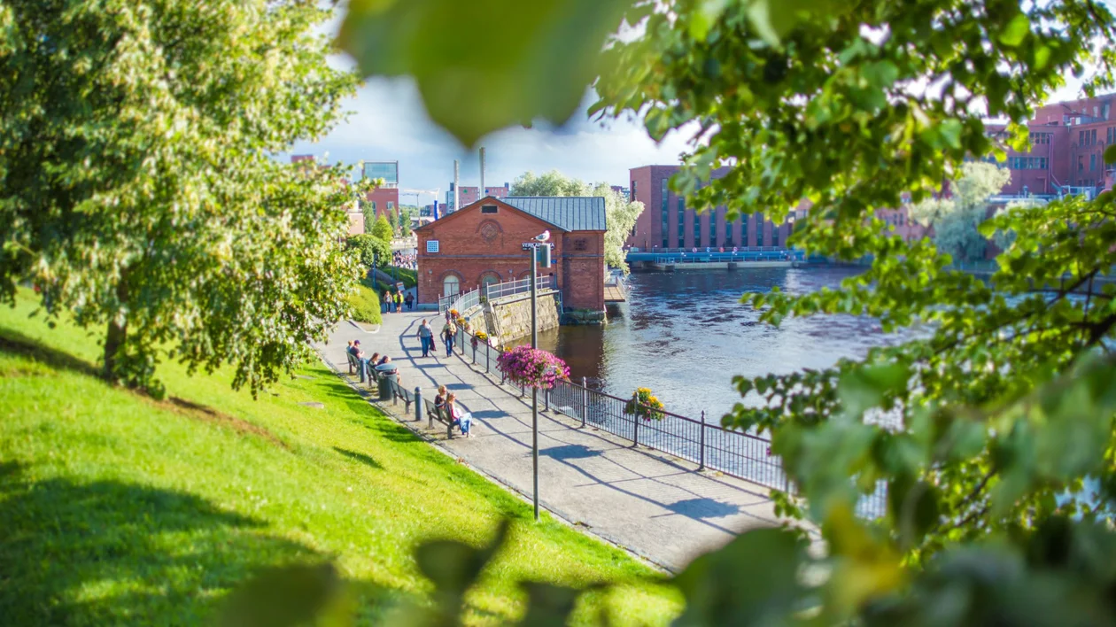 People walking along a riverside path framed by green trees, with red brick buildings nearby.