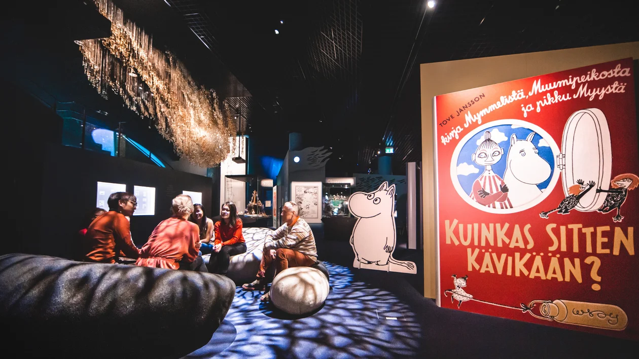 People sitting and talking in a museum with Moomin-themed displays and a colorful poster.