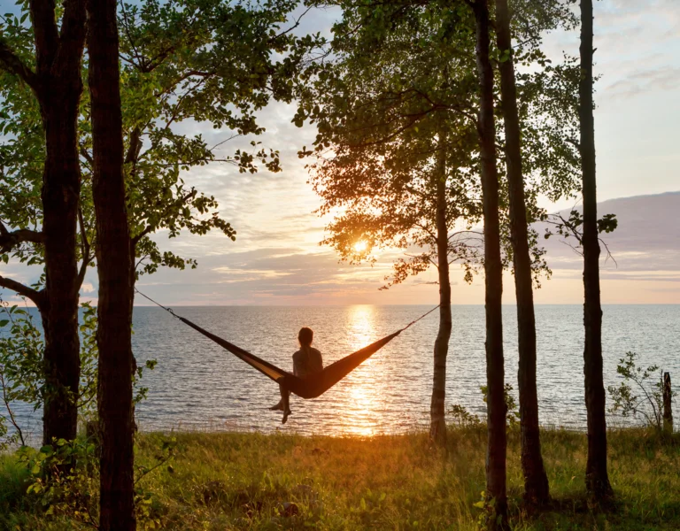 Person relaxing in a hammock between trees at sunset by a lake.