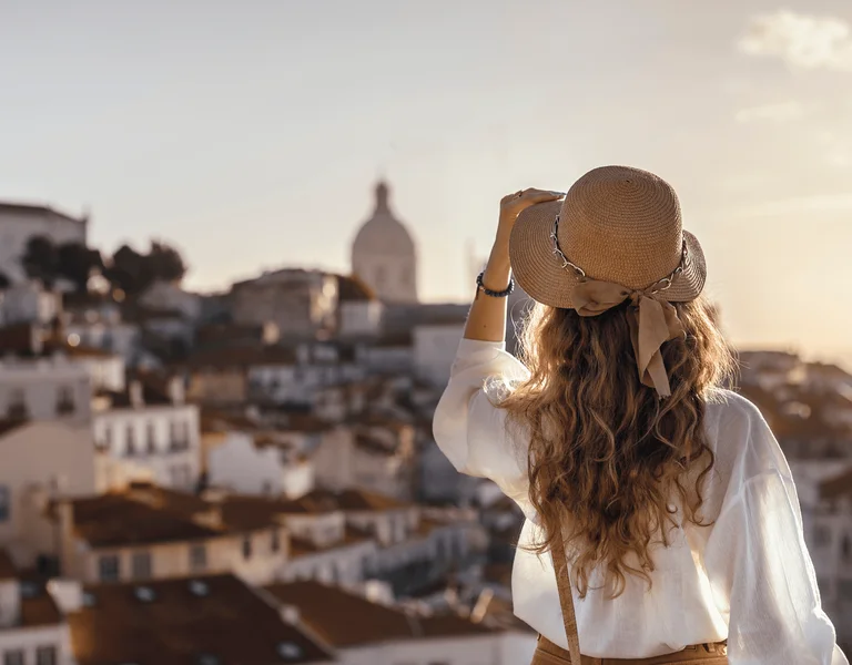 Woman with a hat overlooking sunlit city rooftops and distant dome at sunset.