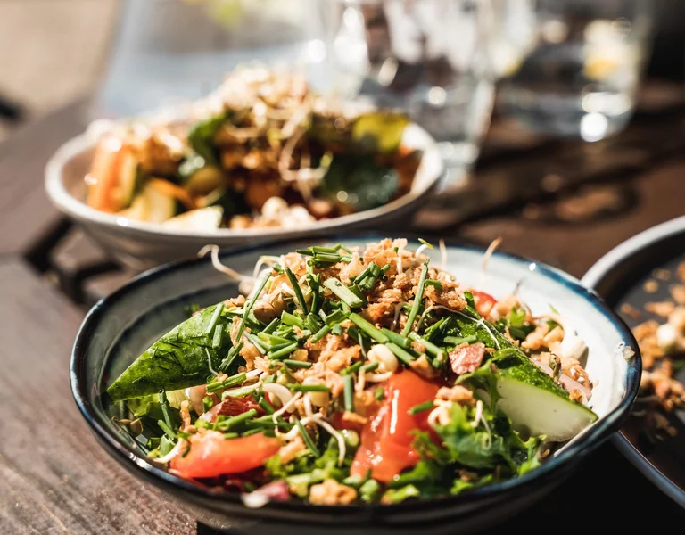 A close-up of a vibrant, fresh salad in a bowl, featuring a mix of leafy greens, cucumbers, tomatoes, sprouts, and crispy fried onions, all garnished with chives. In the background, another salad bowl can be seen, slightly blurred, with the scene lit by natural sunlight, creating a warm and inviting outdoor dining atmosphere.