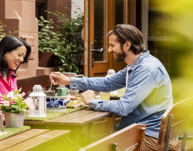 A couple enjoys a cozy outdoor meal at a traditional German restaurant. They are seated at a wooden table adorned with fresh flowers and lanterns, enjoying a relaxed, intimate dining experience surrounded by greenery.